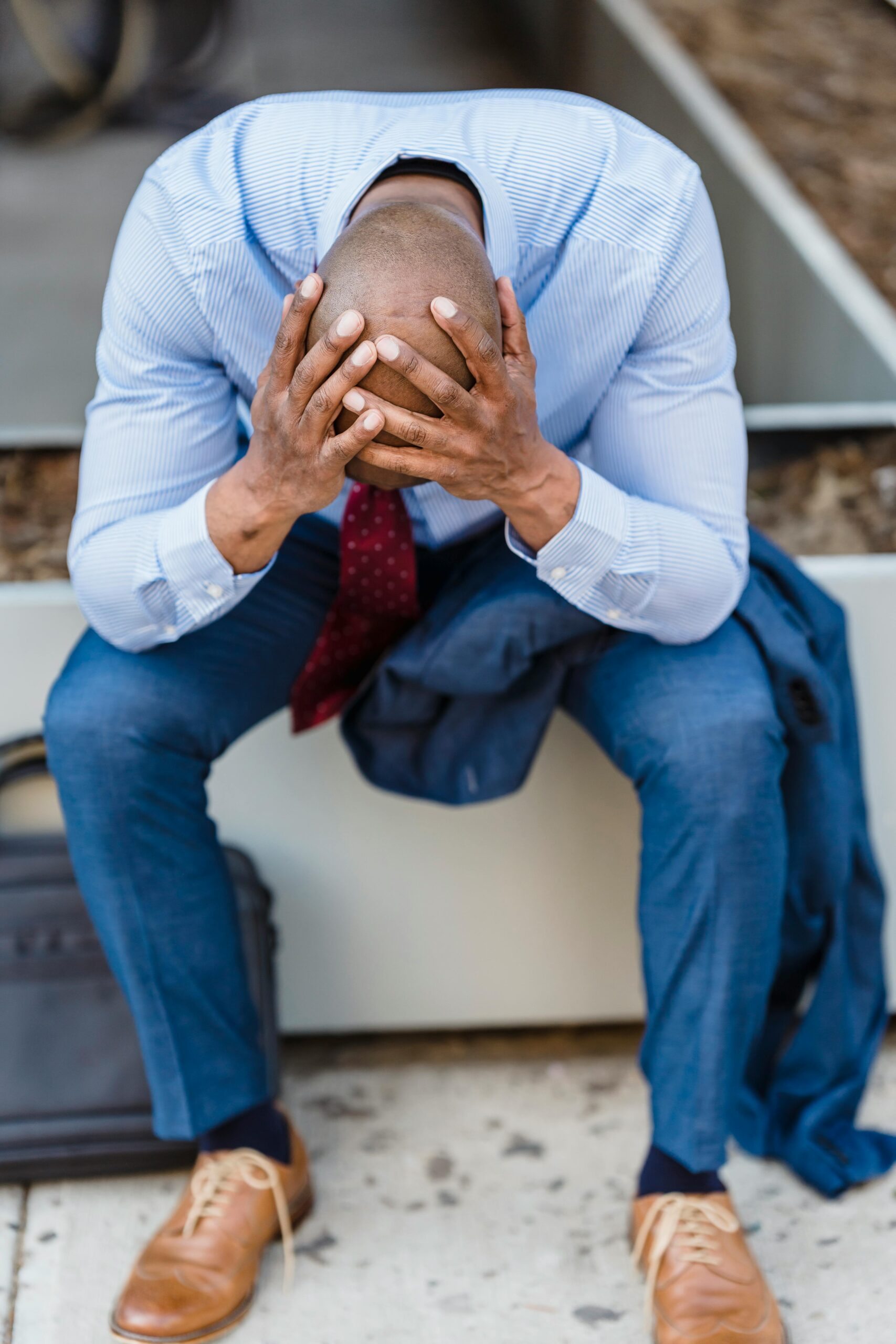 A businessman in a suit sits outdoors, head in hands, expressing stress and despair.